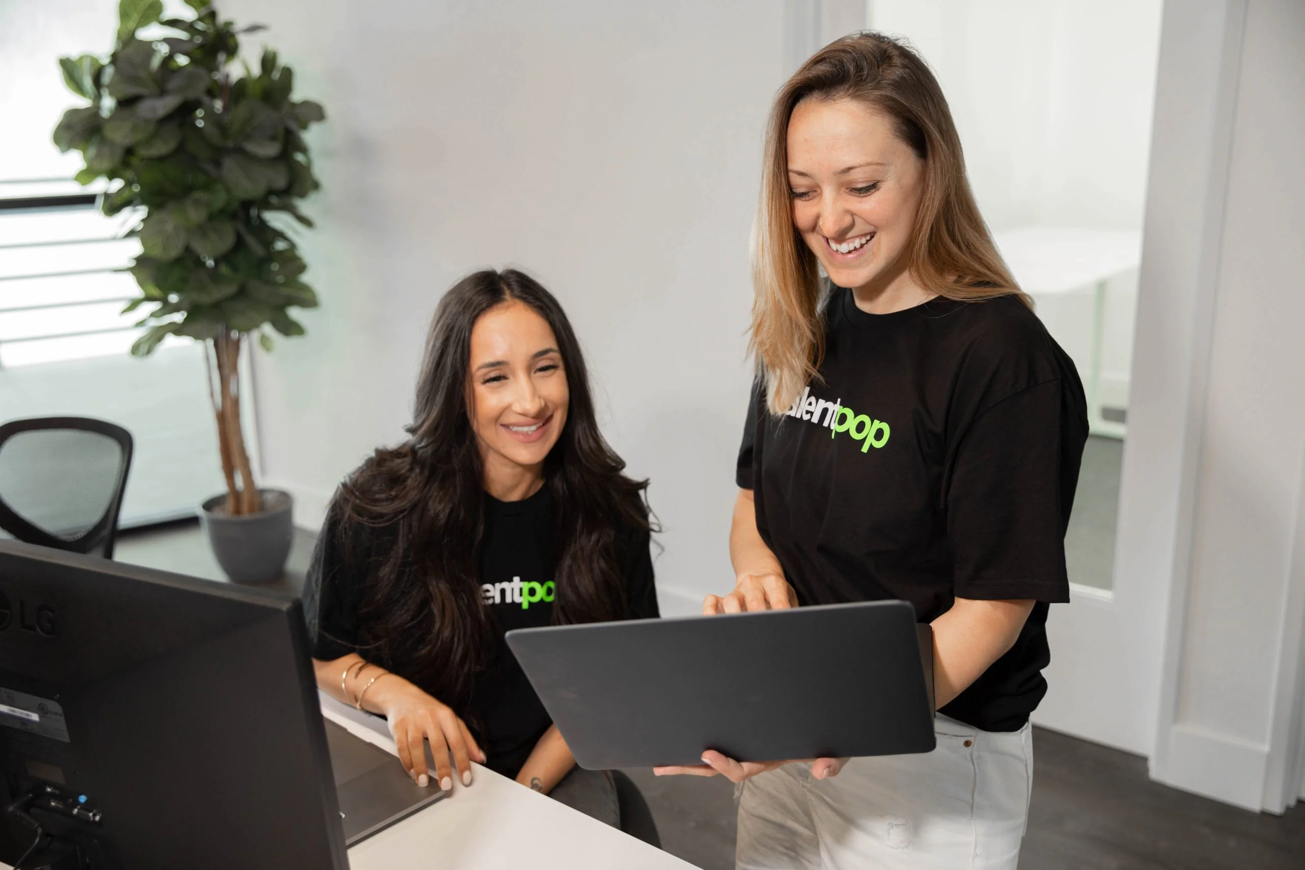 Two women in a modern office setting wearing black t-shirts with the "talentpop" logo in white and lime green; one is seated at a desk with an LG monitor while the other stands beside her holding a laptop.