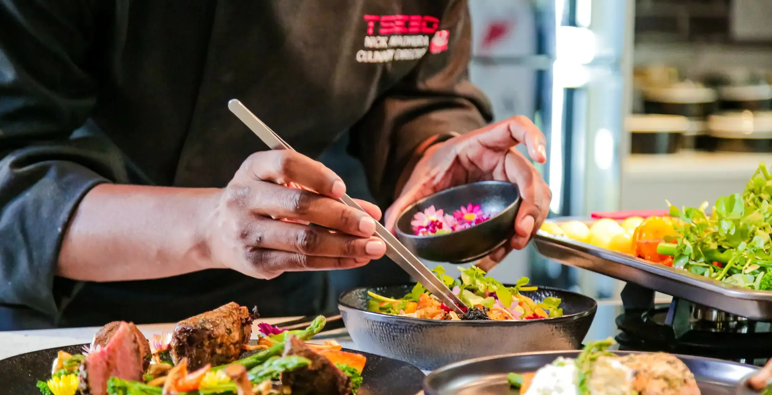 A close-up of a professional chef using tweezers to carefully place edible pink flowers into a black bowl of gourmet salad.