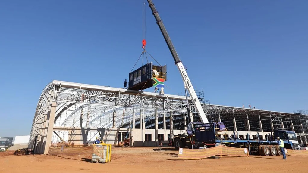 A wide-angle view of an industrial construction site in South Africa. A large red and white mobile crane from ST Crane Hire is lifting a white shipping container or site office. In the background, there is a large industrial warehouse under construction with blue and grey cladding. A white container with a prominent blue and red "MACSTEEL" logo is visible in the foreground.