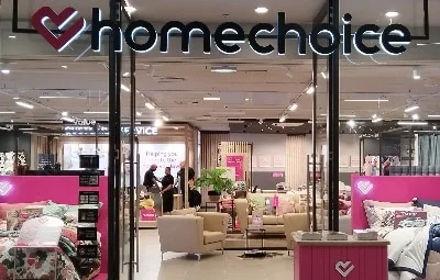 The brightly lit entrance of a HomeChoice store in a mall, featuring pink branding displays, bedding sets, and comfortable lounge furniture.