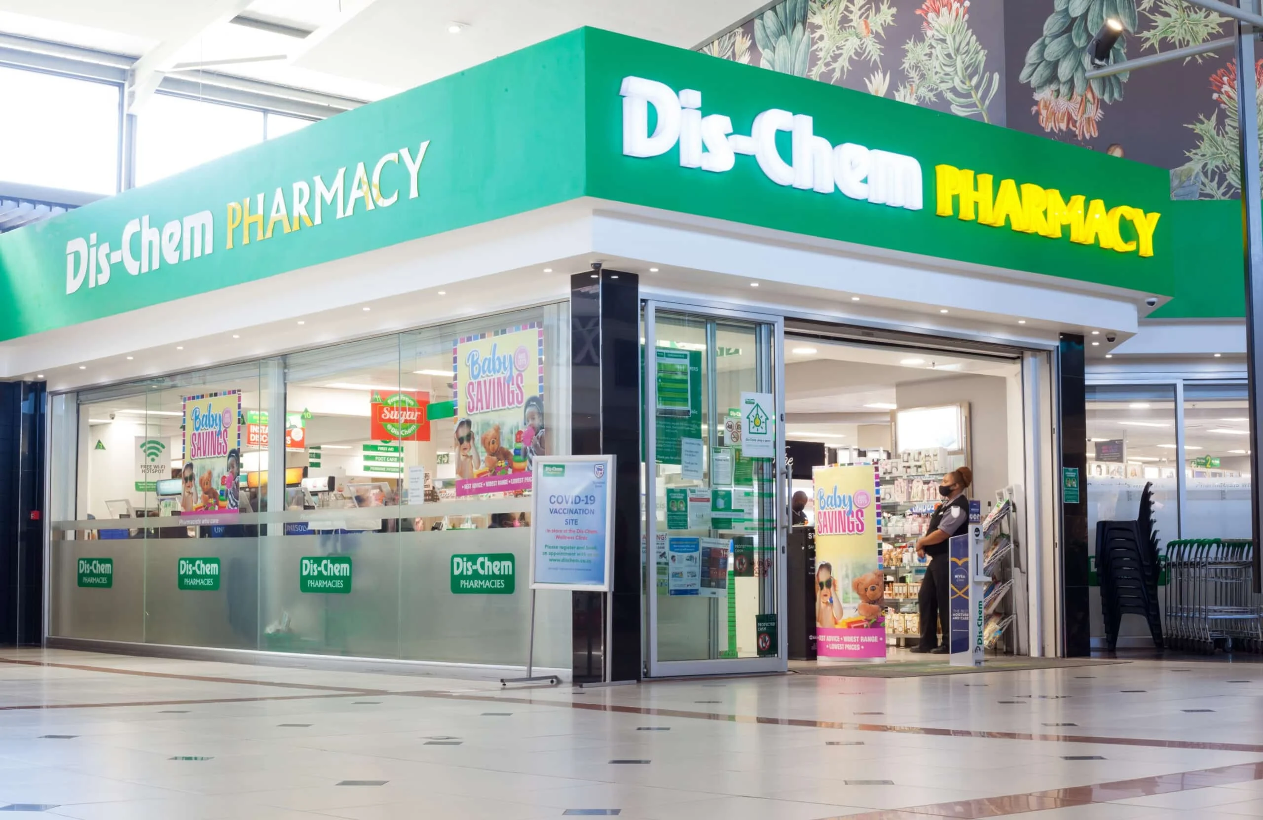 Brightly lit Dis-Chem Pharmacy storefront in a shopping mall, featuring green signage, glass windows with "Baby Savings" posters, and an open entrance with a security guard standing nearby.