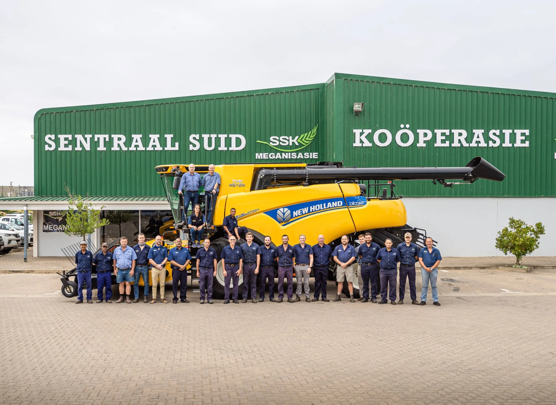 A large group of male and female staff members in blue uniforms standing in front of a giant yellow New Holland combine harvester and a green SSK Meganisasie building.