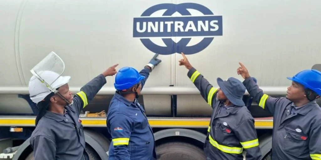 Four industrial workers in navy blue uniforms with yellow reflective stripes and hard hats standing beside a large white Unitrans tanker. They are all pointing towards the prominent blue and white "UNITRANS" logo on the side of the vehicle.