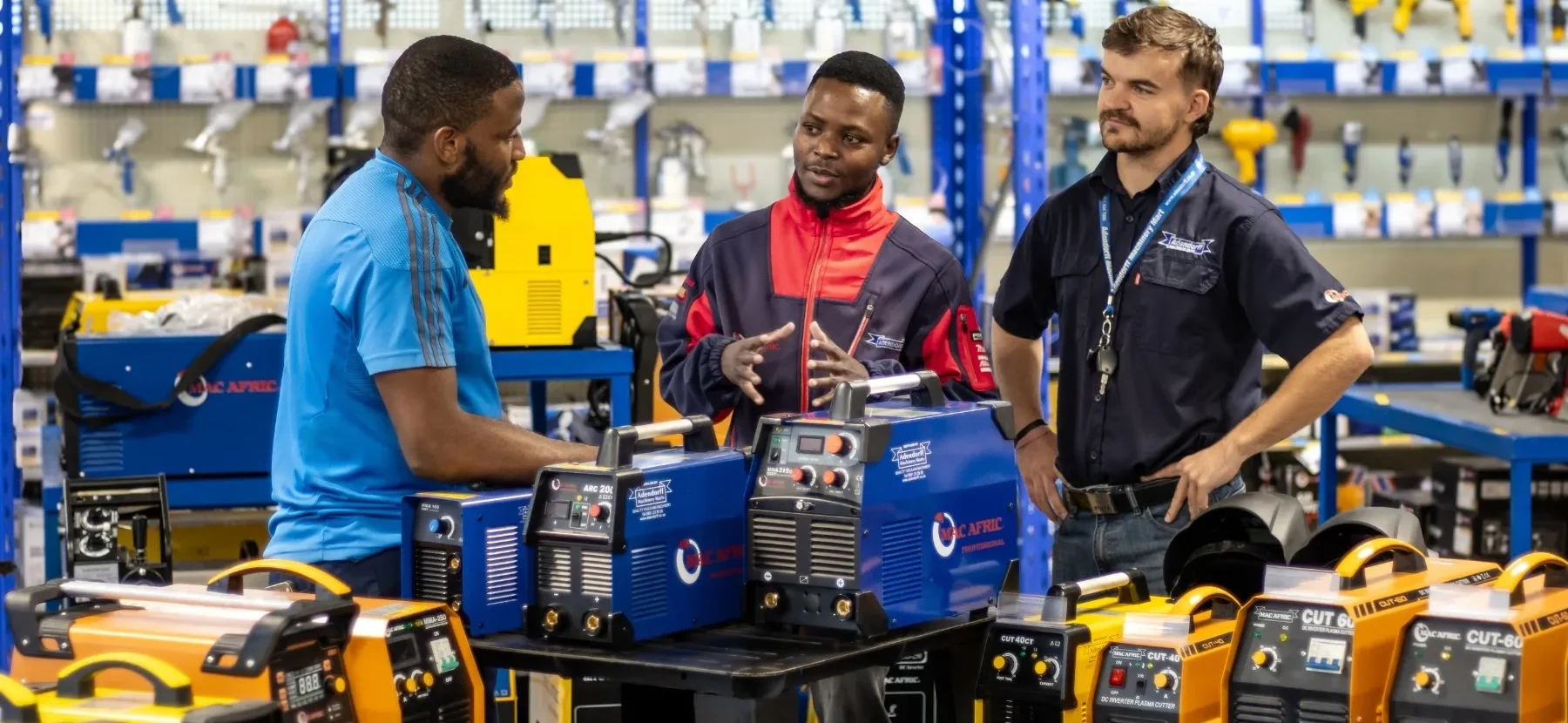 Two staff members in Adendorff branded uniforms providing technical advice to a customer in a tool showroom. They are standing behind a display of blue and yellow MAC AFRIC welding machines, including arc inverters and plasma cutters.