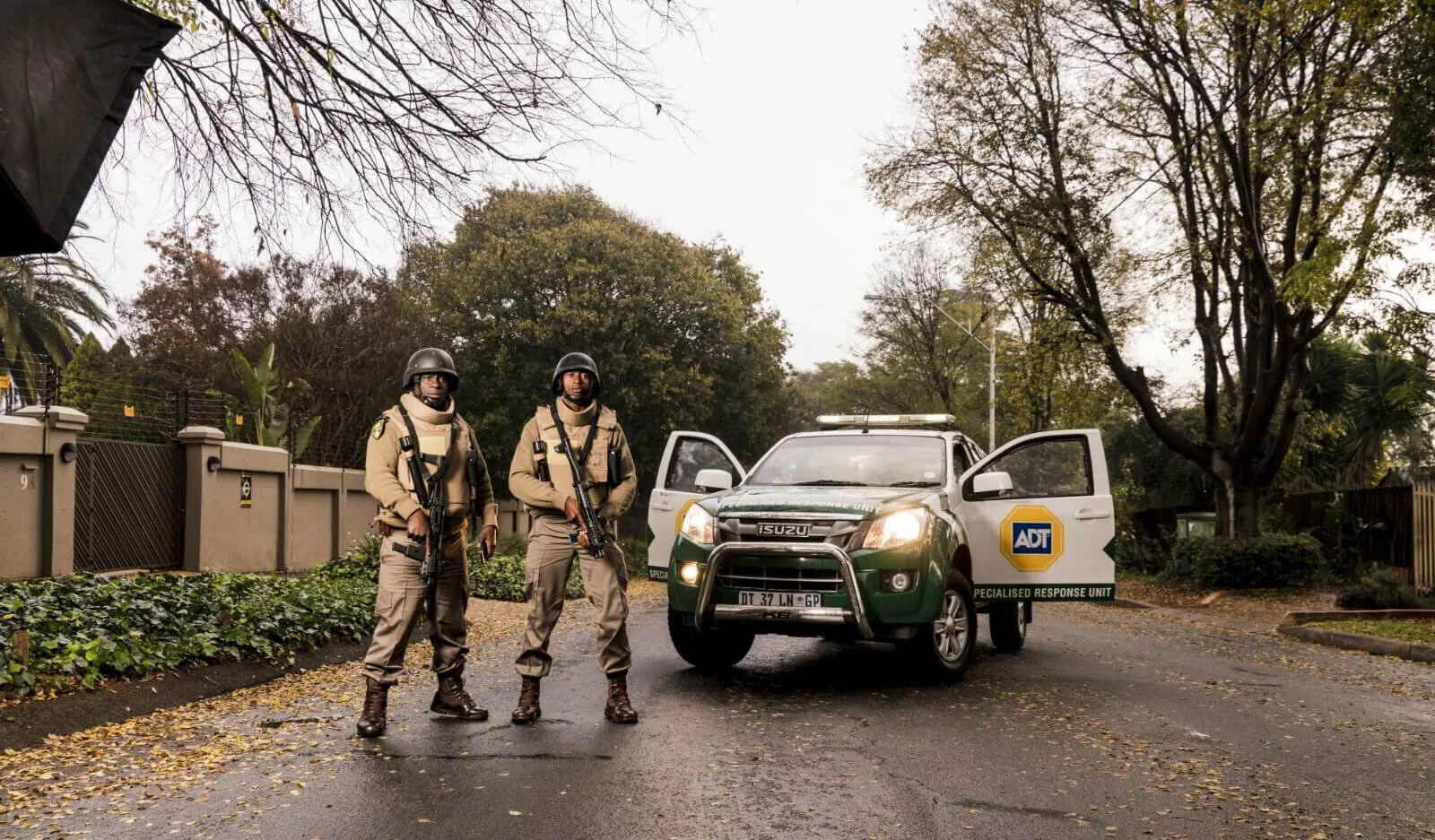 Two security officers in full tactical gear, including helmets and ballistic vests, standing on a leaf-strewn suburban road next to a green and white Isuzu security vehicle. The vehicle features the blue and yellow ADT logo and "SPECIALISED RESPONSE UNIT" branding.