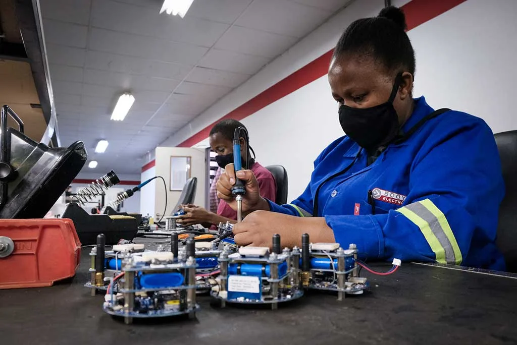 A technician wearing a blue work uniform and a black face mask soldering electronic components at a workbench in a production facility.