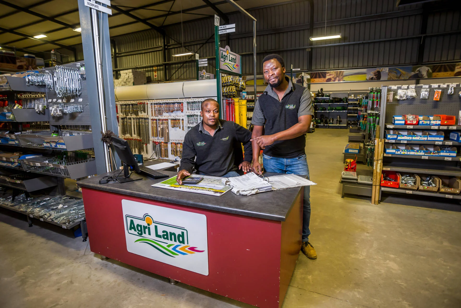 Two retail store employees standing behind an Agri Land service counter inside a hardware and agricultural supply warehouse.
