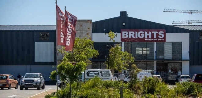 An exterior view of a Brights Hardware store building with large red signage, branded flags, and a parking lot with cars in the foreground.