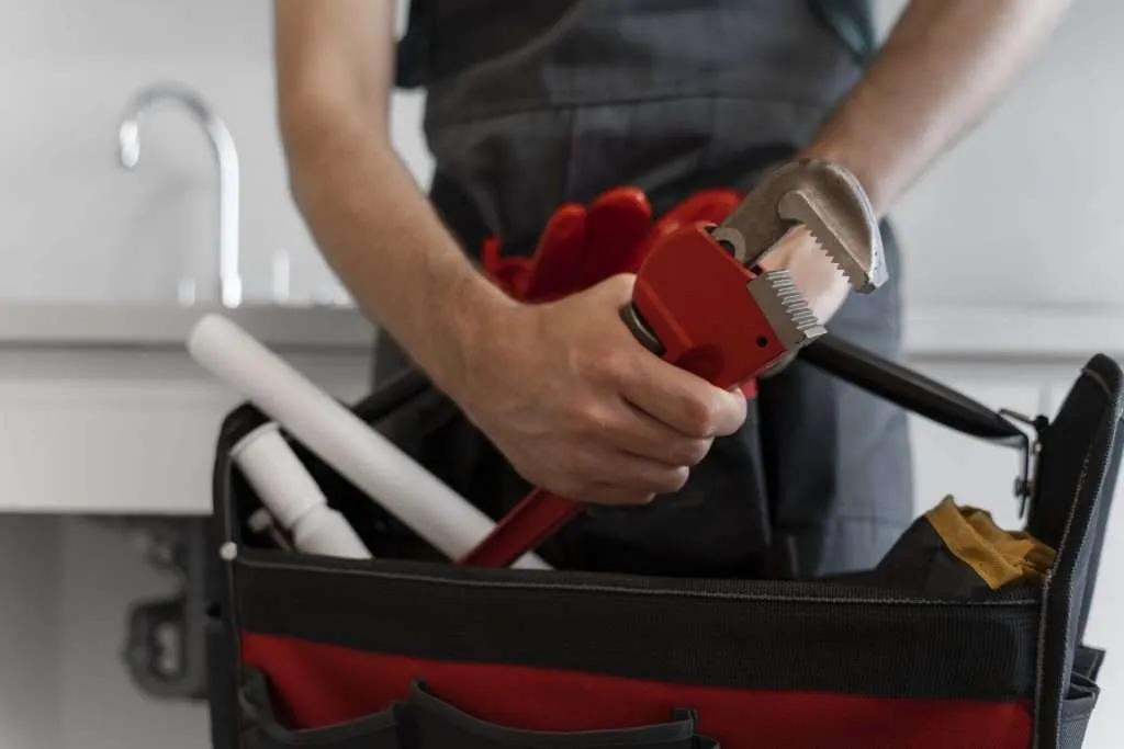 Close-up of a plumber in dark overalls holding a red pipe wrench over a tool bag packed with plumbing supplies.