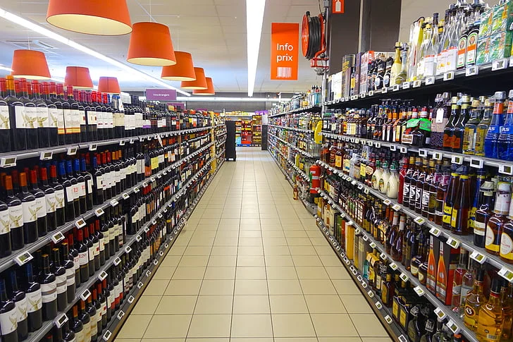 A wide, well-lit supermarket aisle lined with shelves stocked with various wine and liquor bottles under warm hanging lamps.